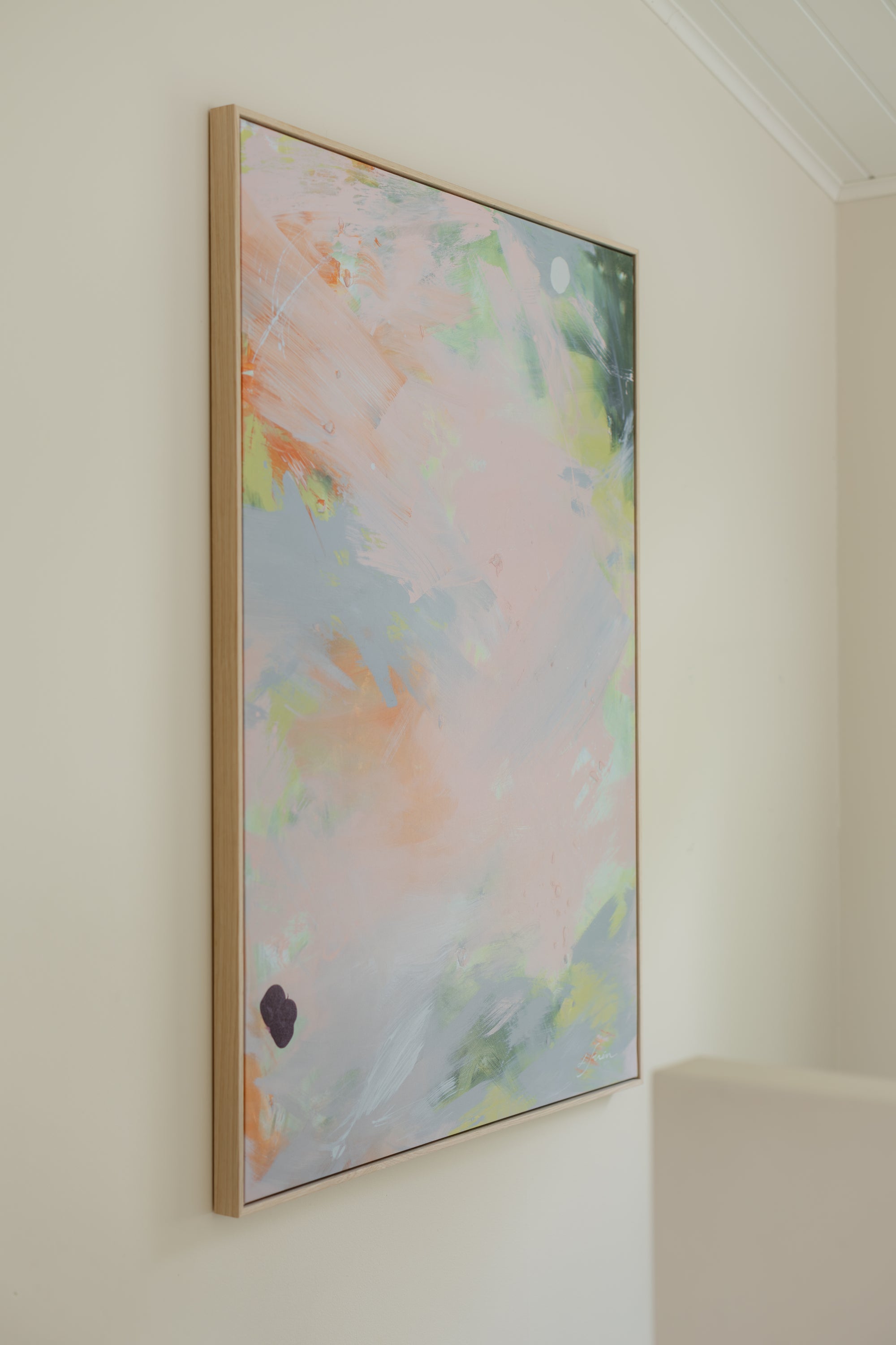 An abstract framed wall art print hanging on a cream-colored wall. The artwork features soft pastel brushstrokes in pink, light blue, green, and peach, enclosed in a thin light wood frame.
