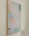 An abstract framed wall art print hanging on a cream-colored wall. The artwork features soft pastel brushstrokes in pink, light blue, green, and peach, enclosed in a thin light wood frame.