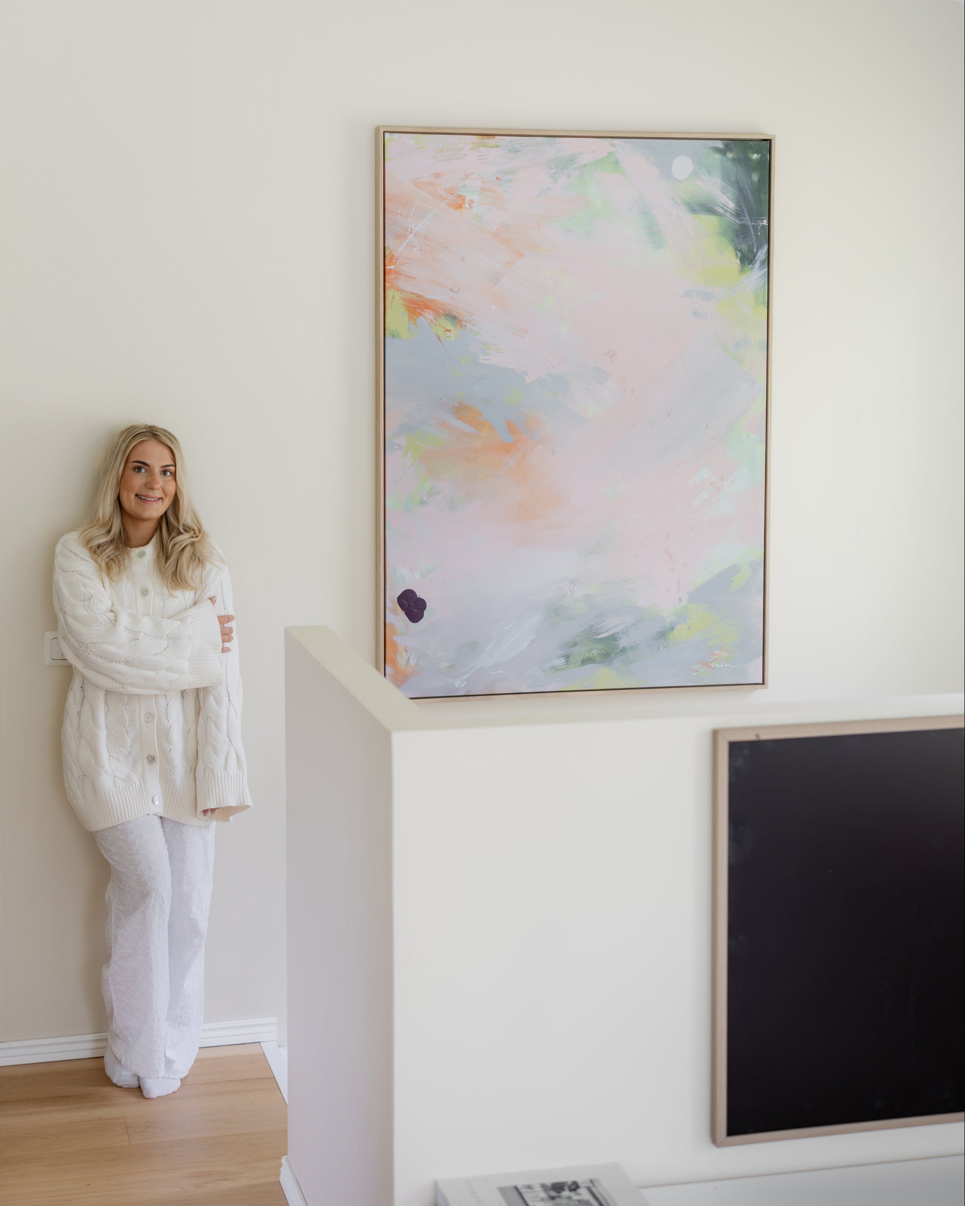 Sara Lindholm in a white knit sweater and white pants stands beside a large framed abstract canvas print  on a white wall in a bright, modern room with light wood flooring.