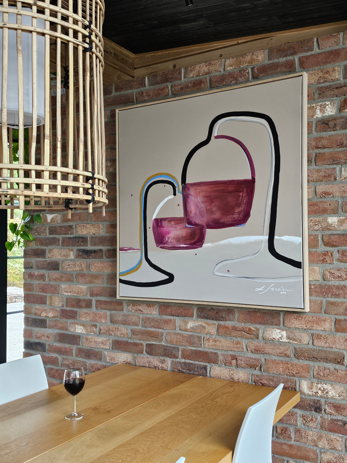 An abstract painting of wine glasses a with pink, purple, black, and blue brushstrokes on a cream background, hanging on a brick wall above a wooden dining table with a glass of red wine.