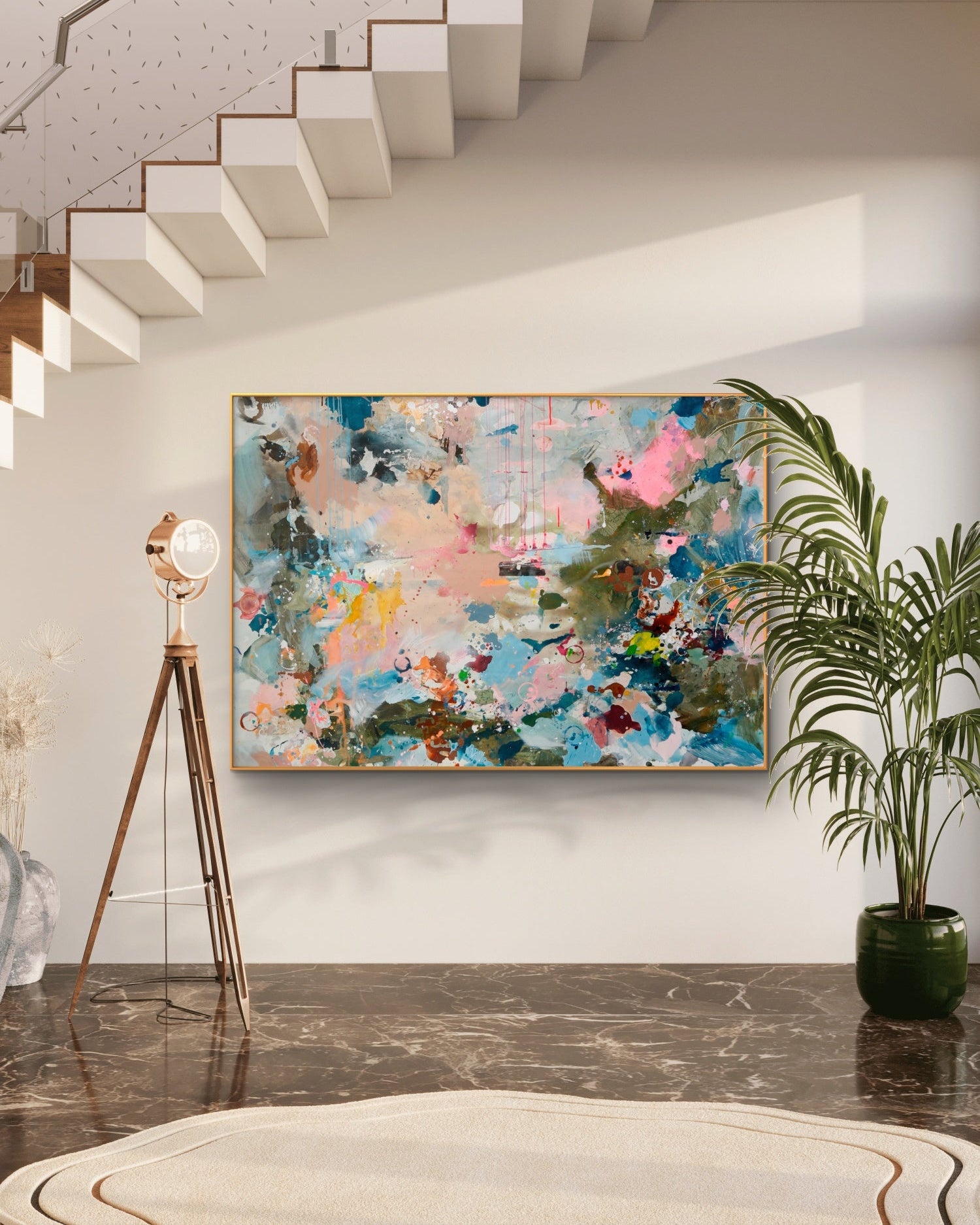 An abstract canvas painting with vibrant splashes of pink, blue, green, and gold, displayed on a white wall in a modern interior setting with a staircase, floor lamp, and potted plant.