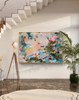 An abstract canvas painting with vibrant splashes of pink, blue, green, and gold, displayed on a white wall in a modern interior setting with a staircase, floor lamp, and potted plant.