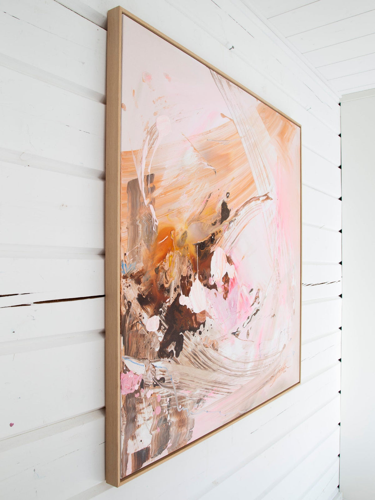 An abstract framed painting with pink, peach, brown, and white brushstrokes, hanging on a white wall.