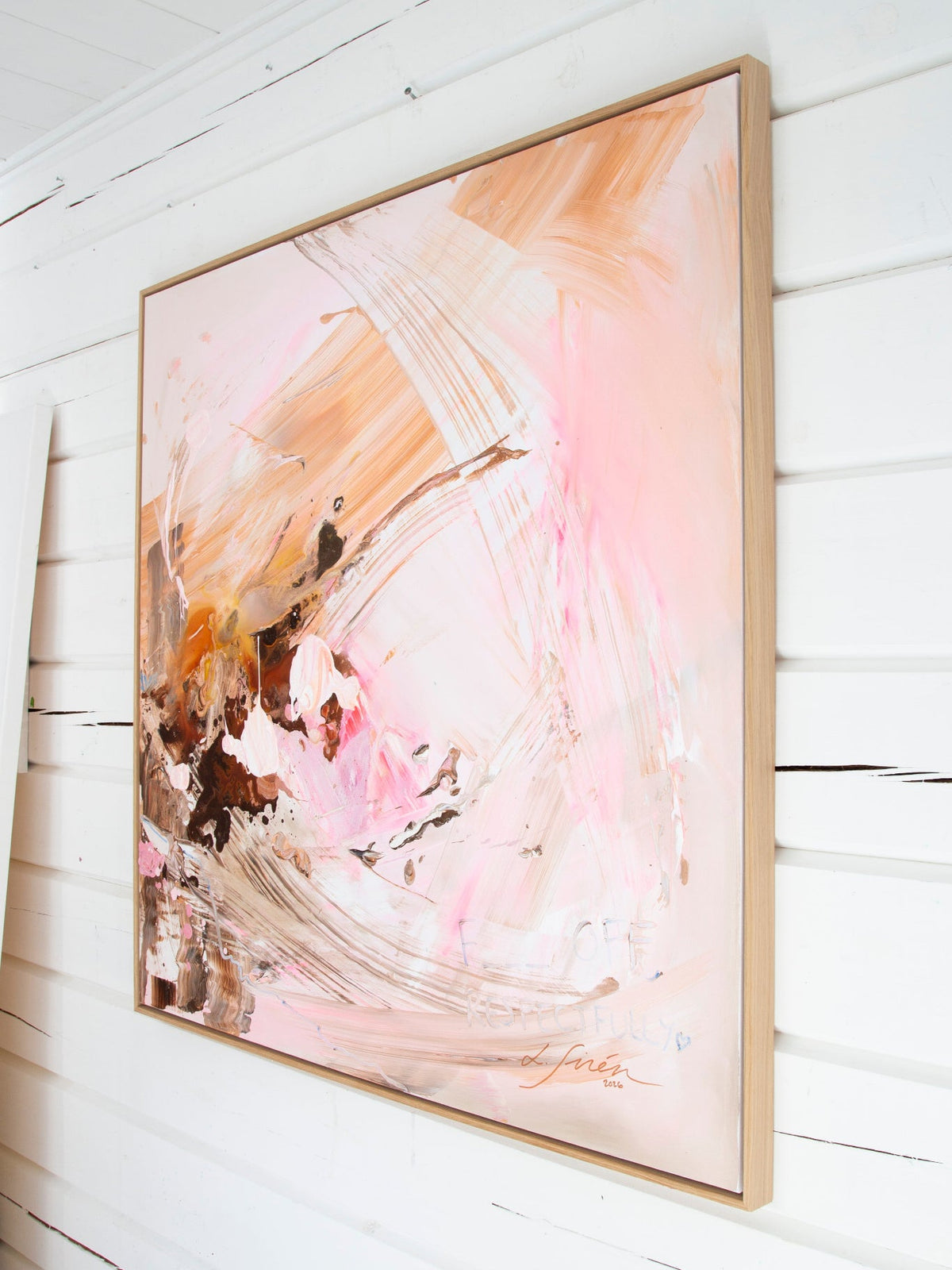 An abstract framed painting with soft pink, peach, and brown brushstrokes, hanging on a white wall.