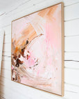 An abstract framed painting with soft pink, peach, and brown brushstrokes, hanging on a white wall.