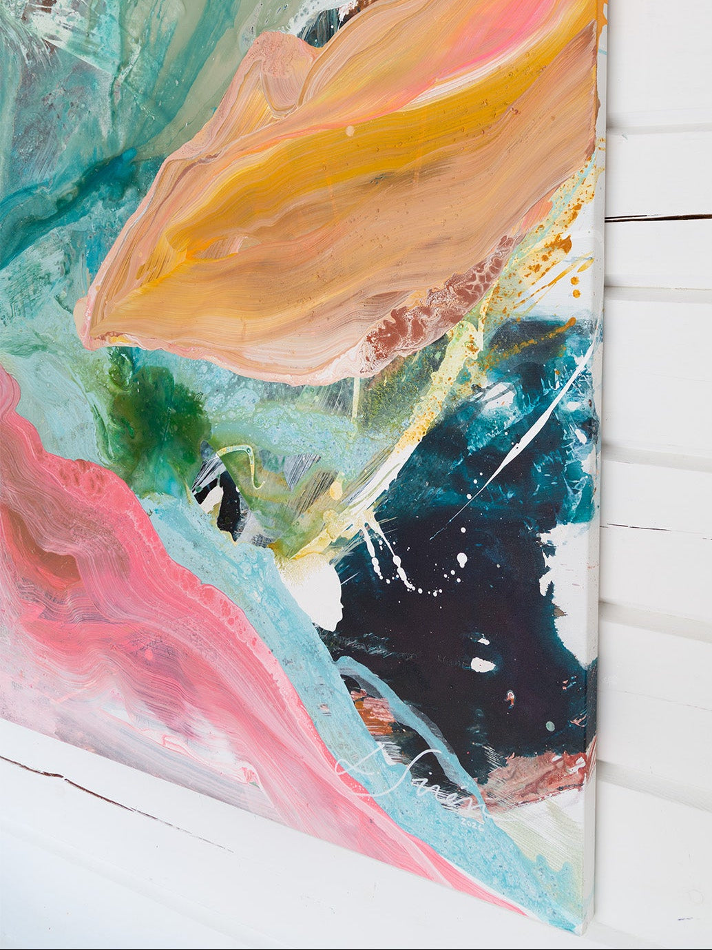 An abstract canvas painting with expressive brushstrokes in teal, pink, peach, orange, black, and white, shown hanging on a white wood-paneled wall.