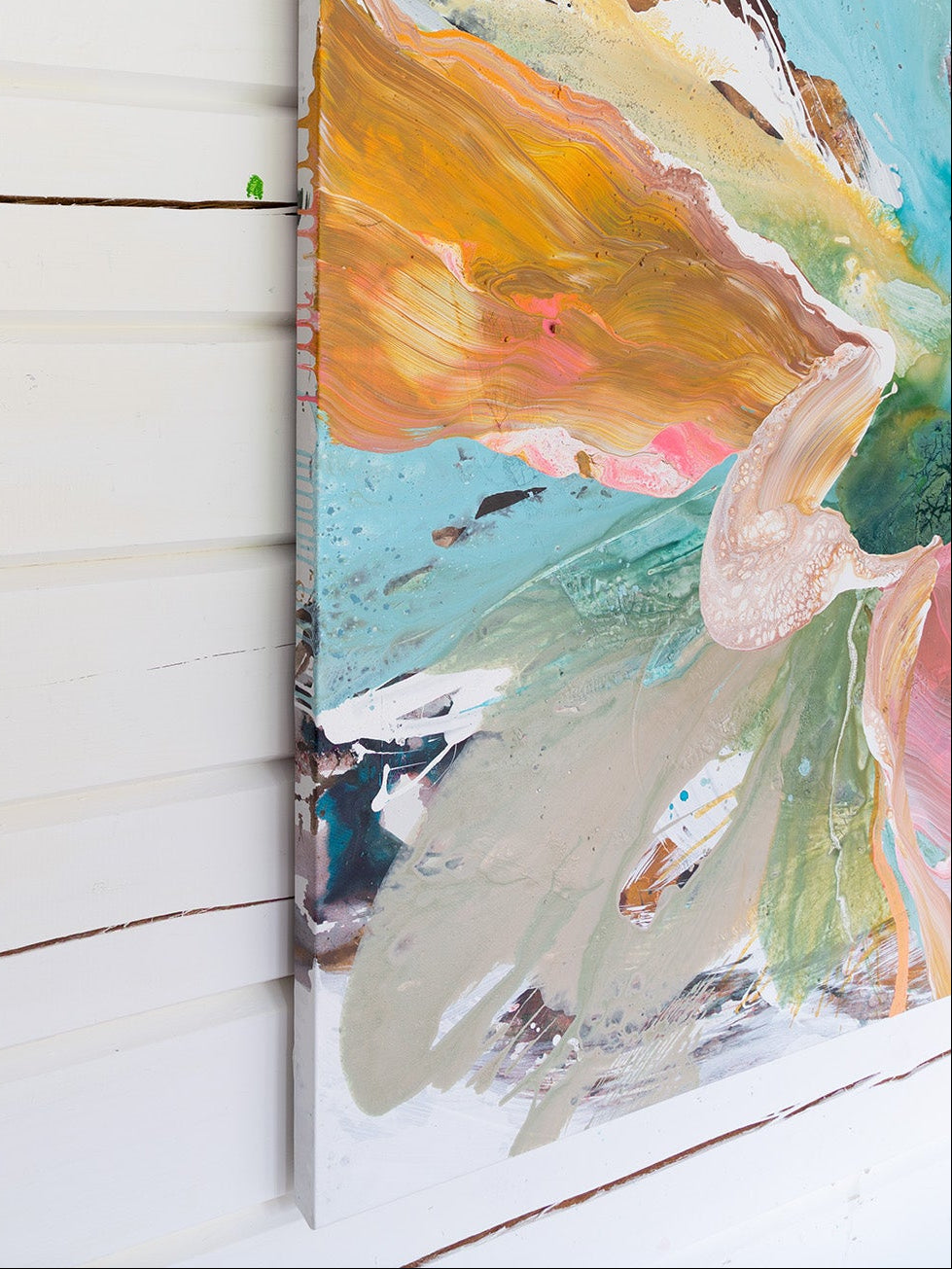 An abstract canvas painting with thick brushstrokes of turquoise, orange, cream, pink, and green, shown hanging against a white wood-paneled wall.