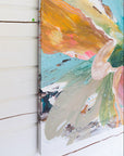 An abstract canvas painting with thick brushstrokes of turquoise, orange, cream, pink, and green, shown hanging against a white wood-paneled wall.