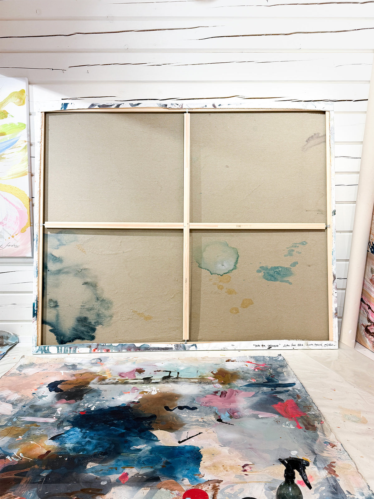 A large four-panel wooden canvas frame   set against a white wood-paneled wall. A paint-splattered palette lies on the floor in the foreground.