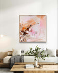An abstract painting with pink, orange, and brown brushstrokes hanging on a white wall above a modern white sofa in a styled living room.