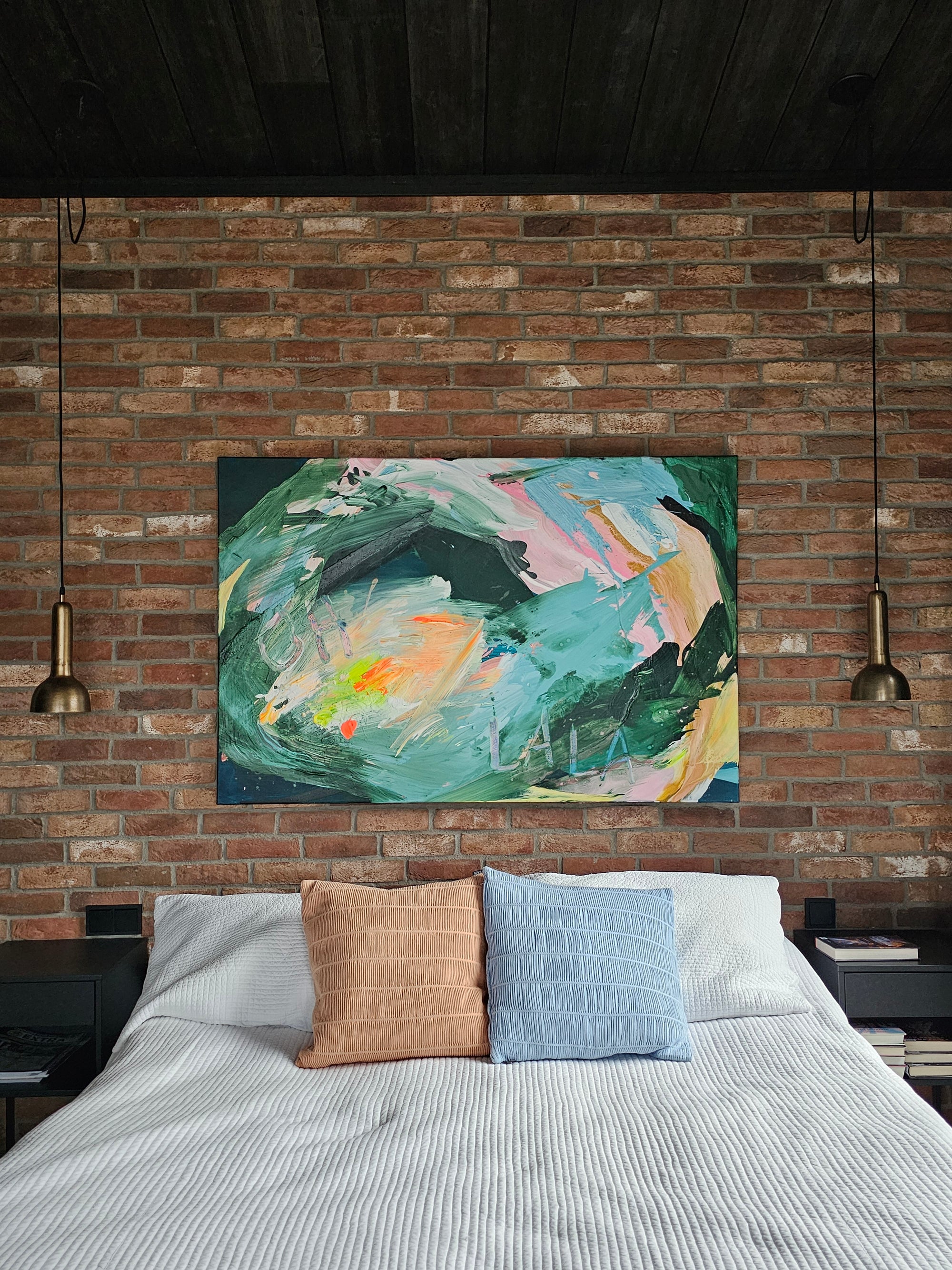 Abstract painting on a brick wall above a bed with colorful pillows.